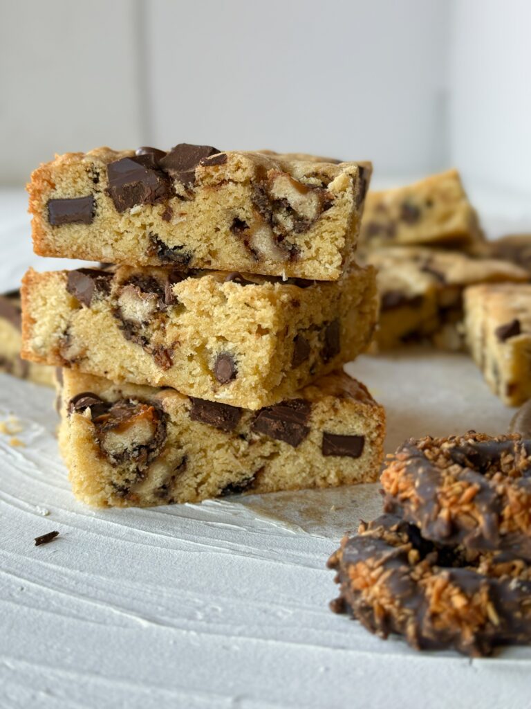 A stack of Samoas Blondies sits on a marble table top with more in the background and a few Samoas in the front of the frame.