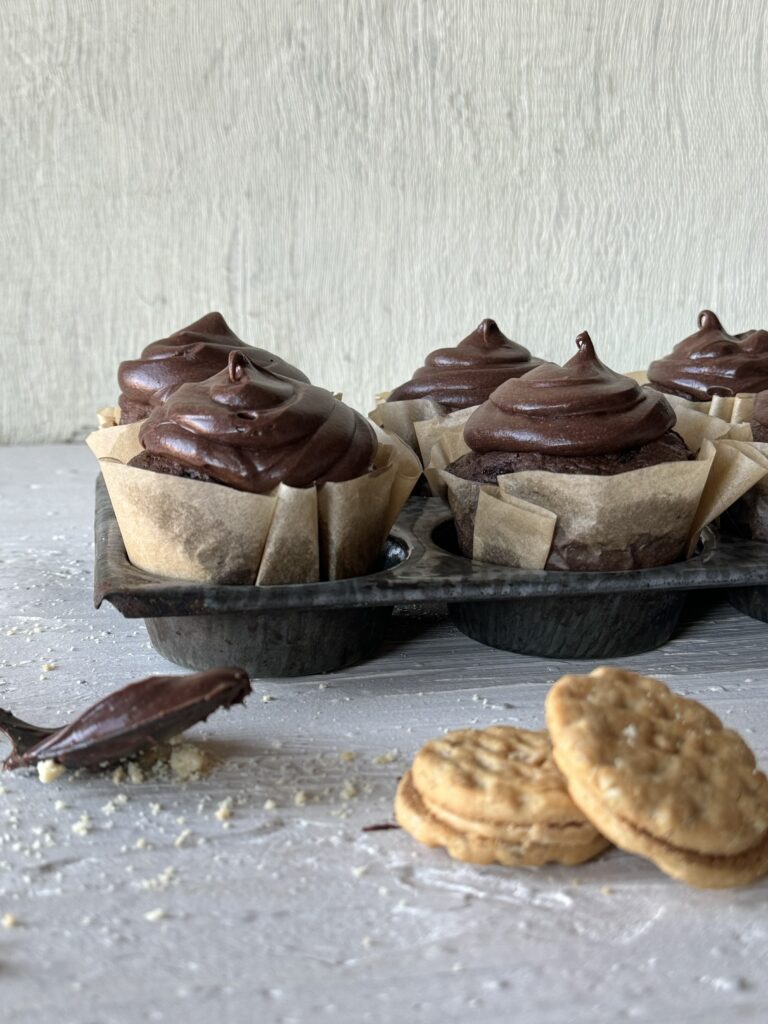 Do-si-dos chocolate cupcakes sit in a muffin tin as a spoon coated in chocolate and two do-si-dos cookies sit in front of the tray.