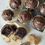 A gathering of chocolate cupcakes with chocolate frosting as one sits split open in front of the group and two do-si-dos cookies sit to the right next to a spoon.