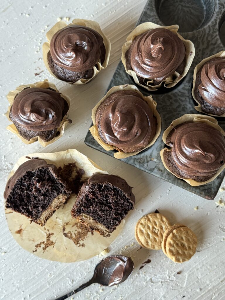 A gathering of chocolate cupcakes with chocolate frosting as one sits split open in front of the group and two do-si-dos cookies sit to the right next to a spoon.
