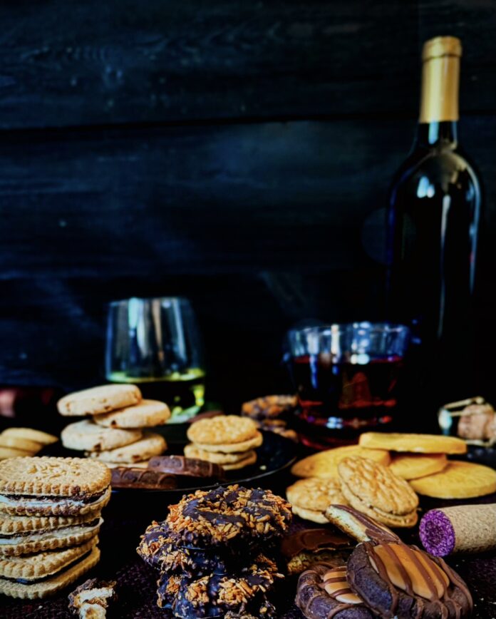 A look in photo of piles of Girl Scout cookies with a bottle of red wine and glasses of white and red wine and wine corks with a black background.