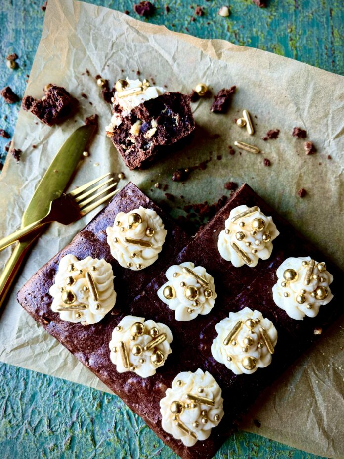 Guinness Brownies with Bailey Buttercream Icing An overhead shot of brownies with buttercream icing and gold sprinkles on a piece of parchment paper on a green surface.