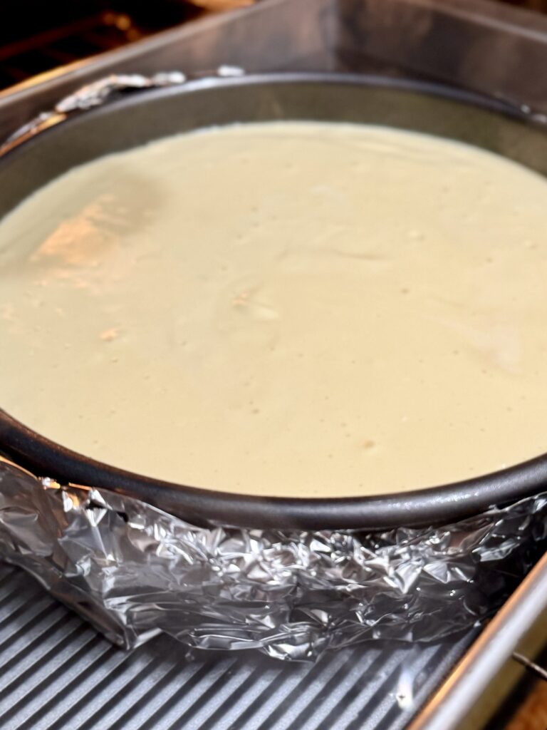 A raw cheesecake with foil wrapped around the bottom and up the sides, sitting in a water bath in a silver baking pan being placed into an oven.