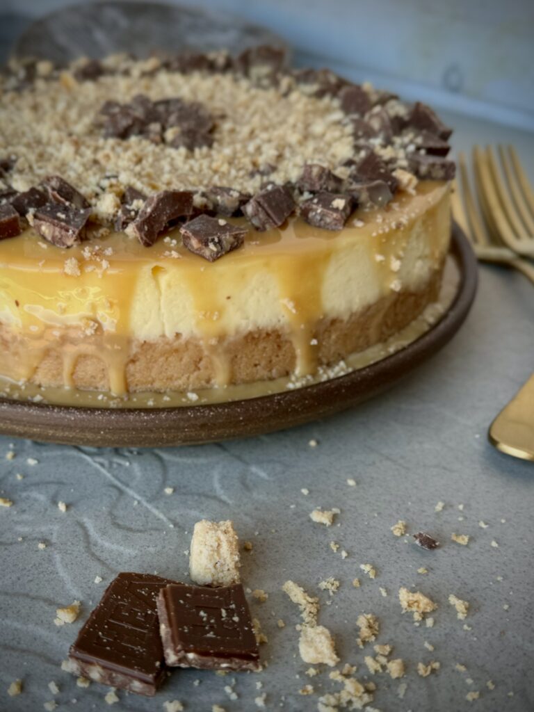 A look-in photo of a cheesecake with caramel dripping down the sides, garnished with chocolate pieces.