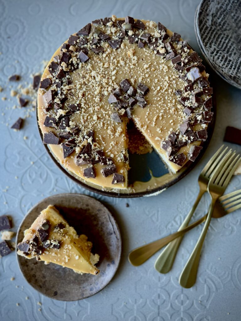An overhead picture of a cheesecake with caramel and chocolate pieced around the edges, and a slice of the cheesecake on a plate with 3 gold forks.