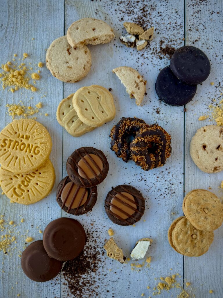 To represent Girl Scout Cookie Inspired Recipes, various Girl Scout Cookies surrounding a panel table and some crumbled up into crumbs.