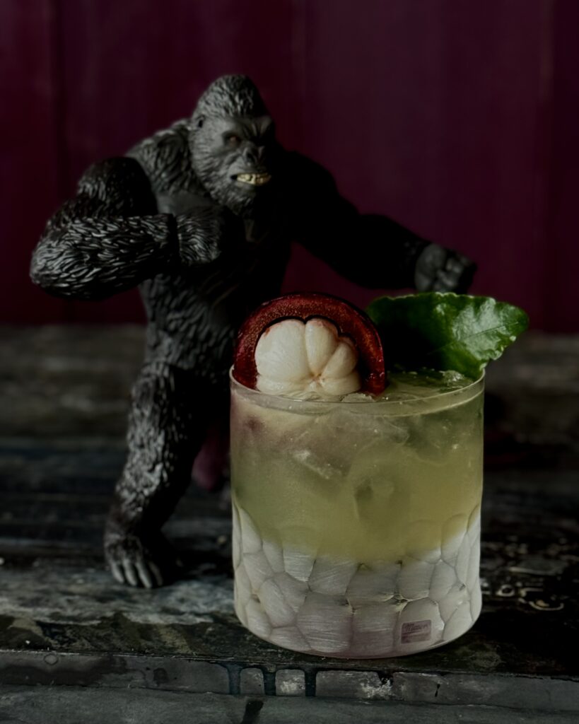 A photo of a light yellow cocktail in a fancy rocks glass with a King Kong figurine next to the glass on a peeling black surface with a deep purple background