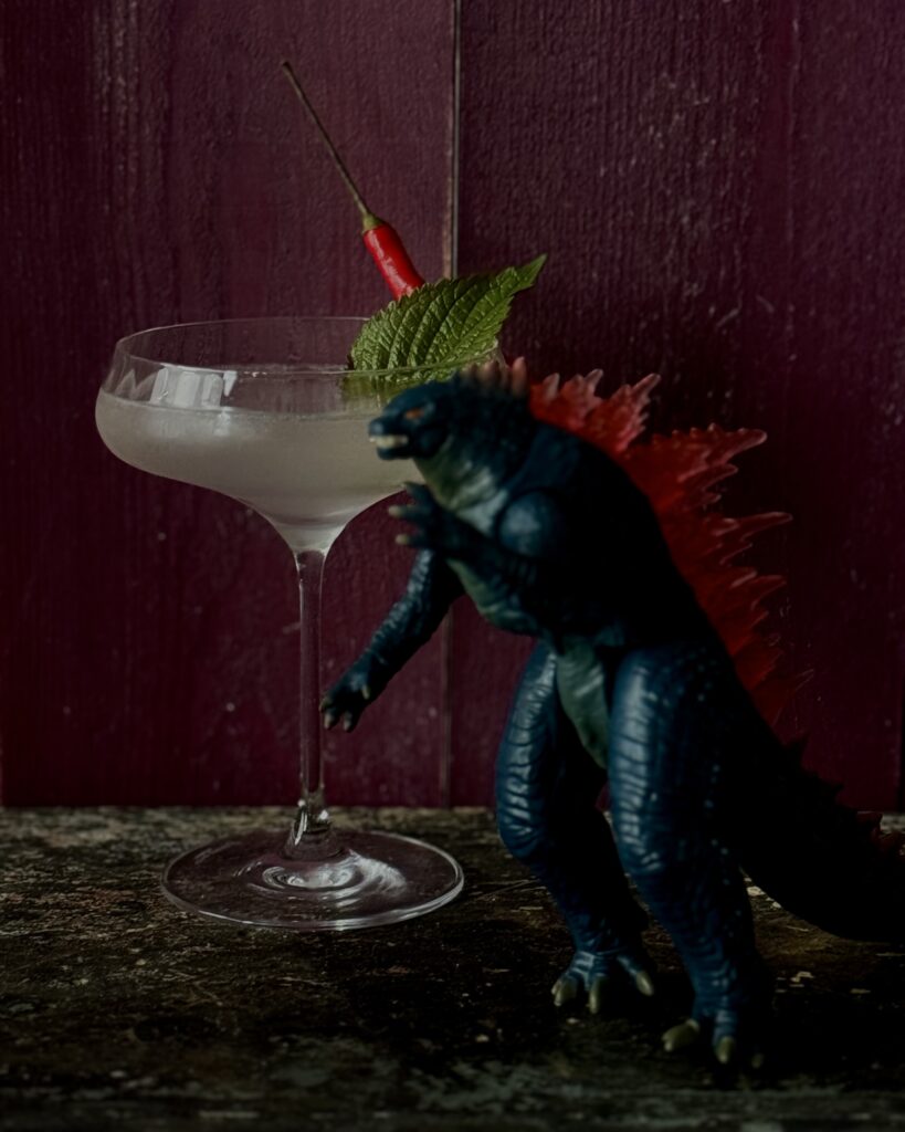 A coupe glass with a light colored cocktail garnished with a red pepper and shiso leaves with a blurred. Godzilla figurine in front of the cocktail