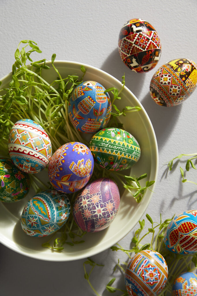 A bowl holds Pysanky designed Ukrainian Easter eggs with green easter grass underneath them and some loose on the table.