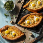 Bread in the shape of pointed ovals and filled with eggs sit on a table with herbs around them. Kachapuri recipe