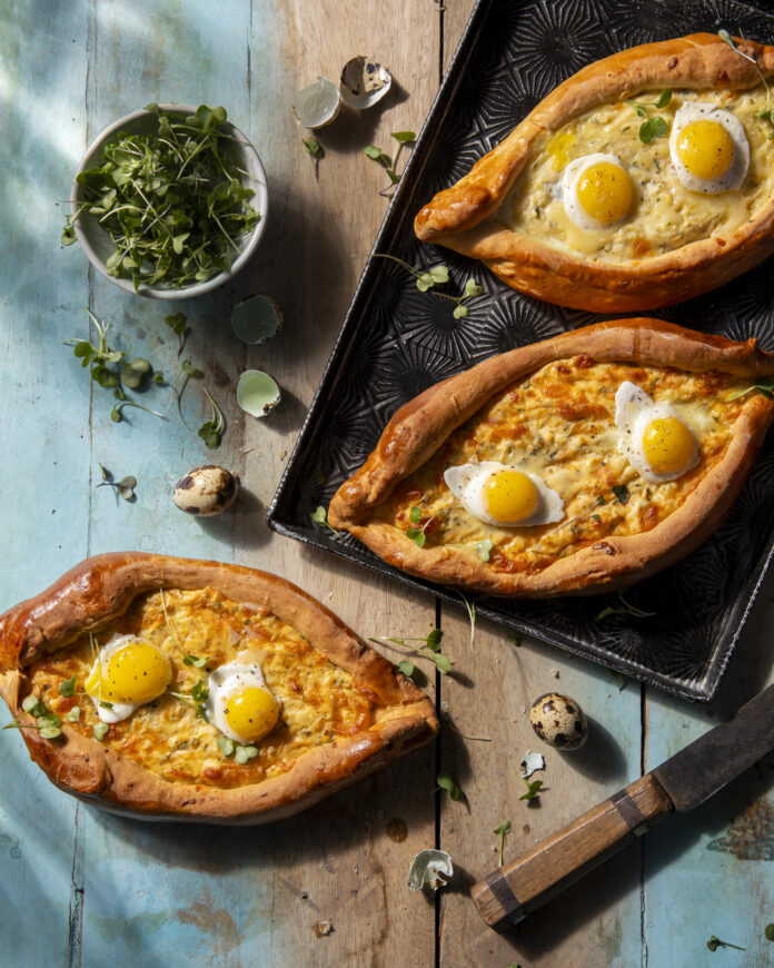 Bread in the shape of pointed ovals and filled with eggs sit on a table with herbs around them. Kachapuri recipe