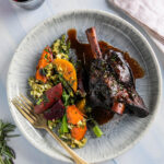 A white plate holds a lamb dinner with burgundy braised lamb shank, roasted vegetables, and a homemade dressing, all on a white table.
