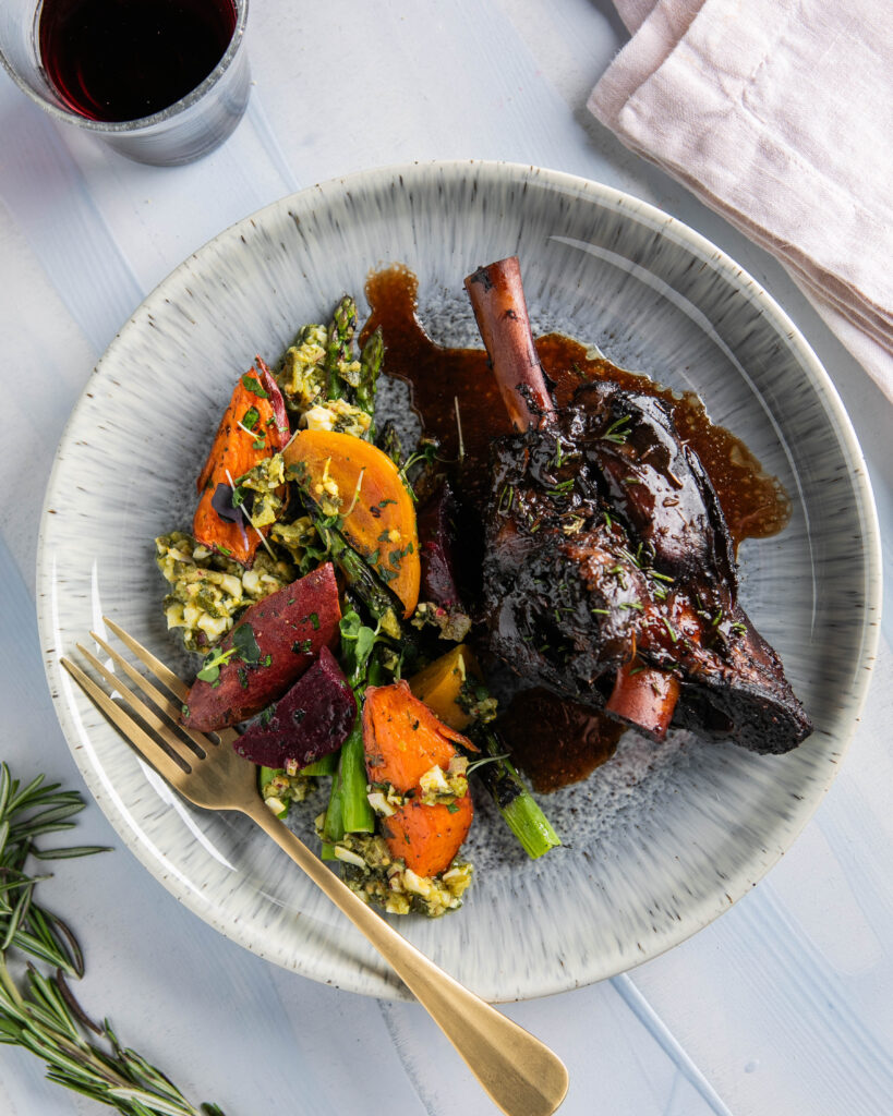 A white plate holds a lamb dinner with burgundy braised lamb shank, roasted vegetables, and a homemade dressing, all on a white table.