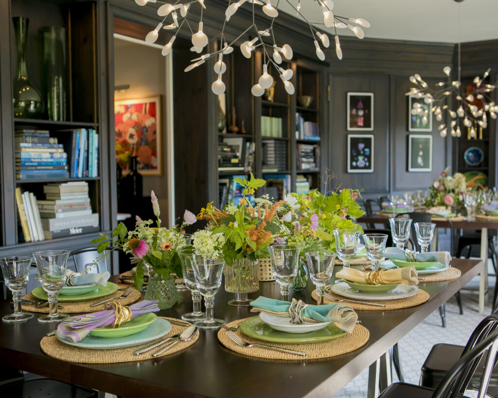 The interior of a darkly painted dining room sits a dining table decorated with brown placemats, lots of wild flowers, and blue plates.