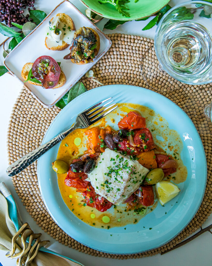 A light blue plate holds a dish of tomatoes in a sauce with a fork positioned at the top of the plate. On the placemat sits anotehr small plate and a glass of water.