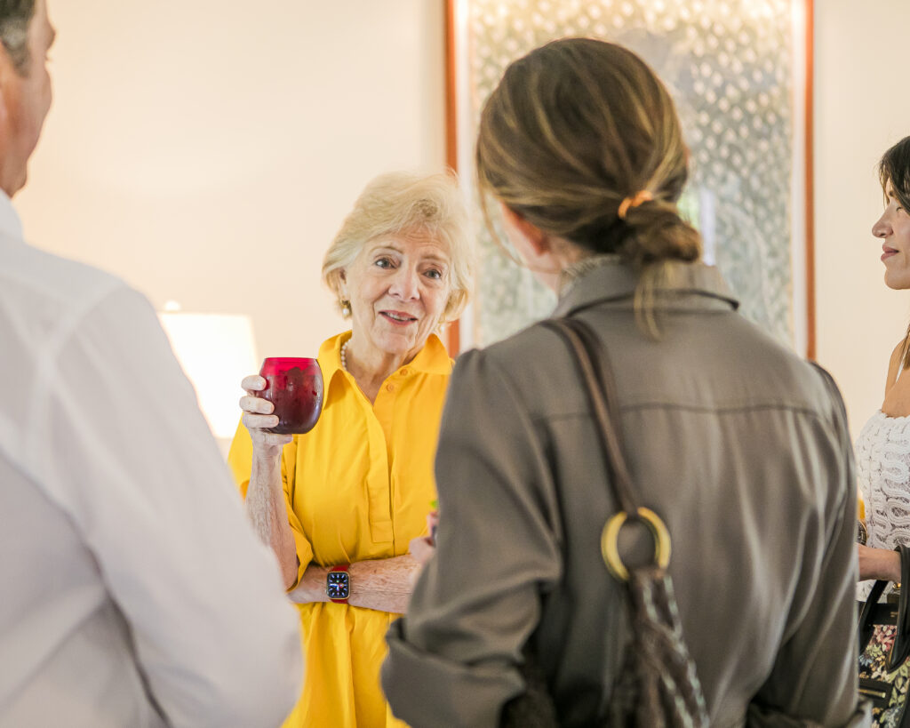 Susan Gravely in a yellow dress holds a red glass in her hand and talks to two people whose backs are towards the camera.