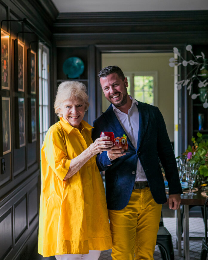 A man in yellow pants and a blue jacket and a woman in a yellow dress cheers their glasses in the entryway of a home.