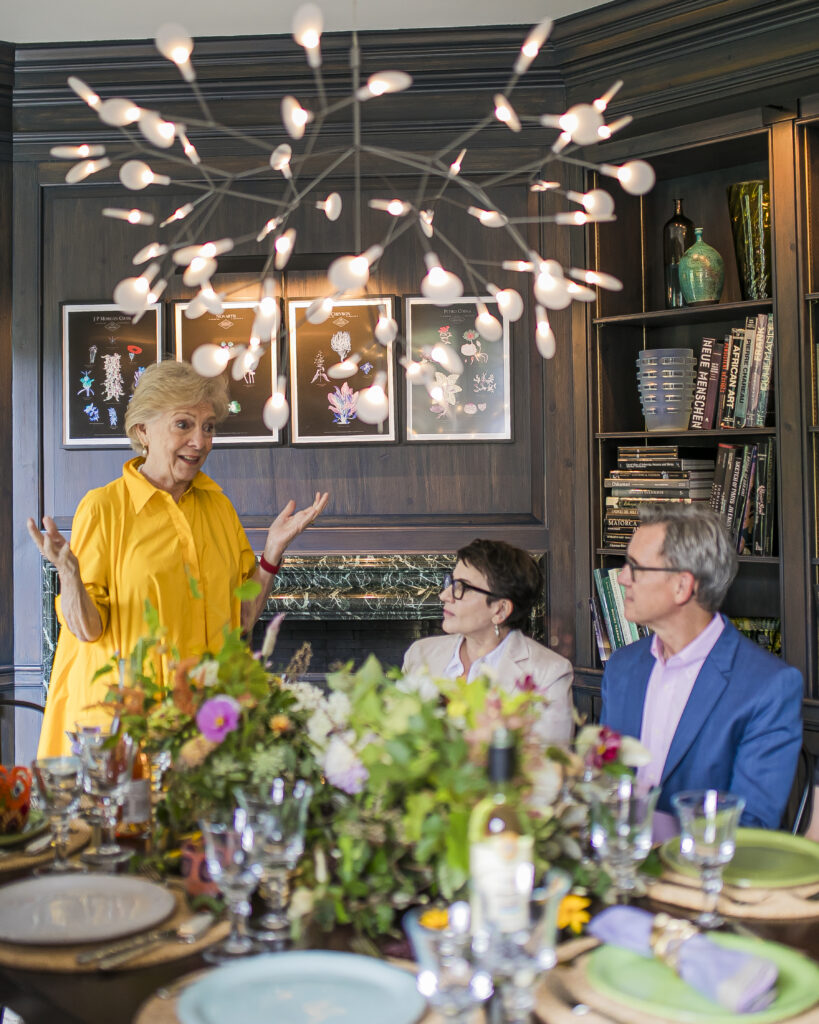 Susan Gravely in a yellow dress speaks to a man and woman seated at the dining table with a fancy light fixture over head.