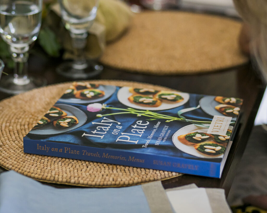 The book Italy on a Plate by Susan Gravely sits on a brown placemat of a dining table.