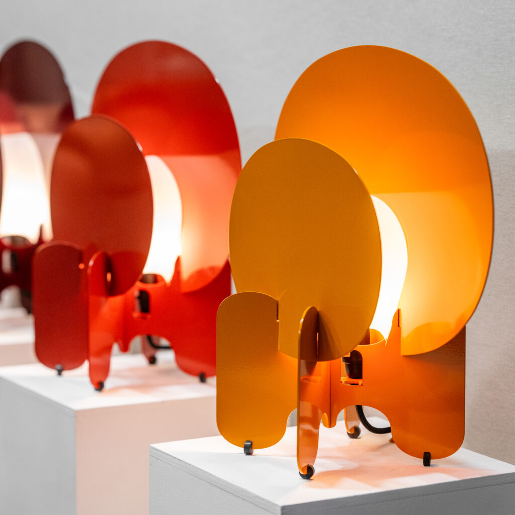 Oval shaped table lamps in the colors orange and red sit on white podiums, lit up between their two ovals.