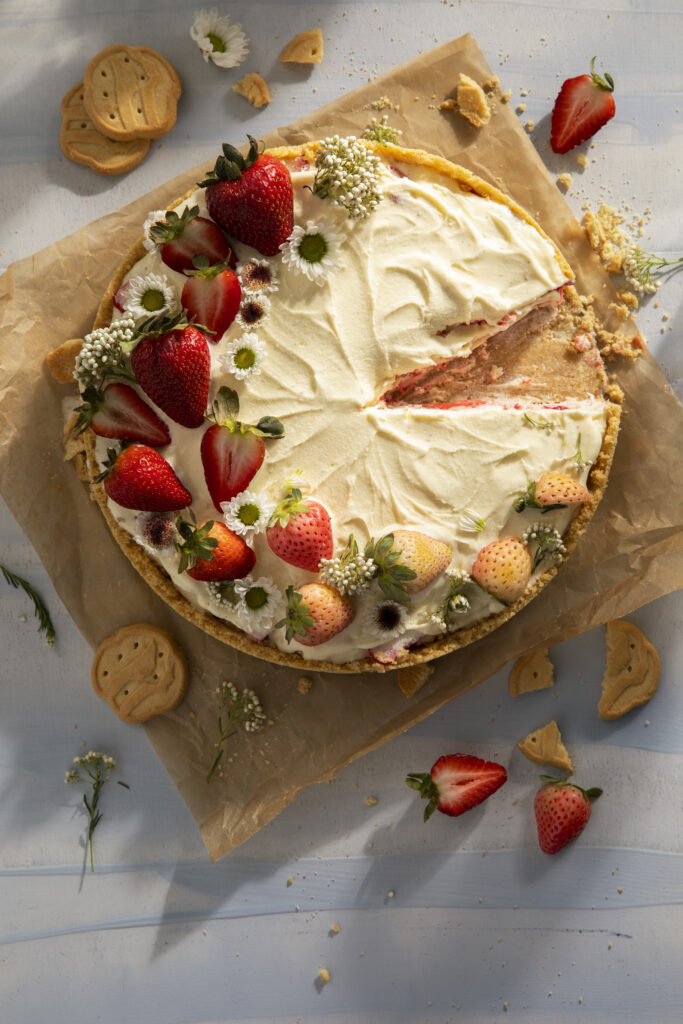 An ice cream cake pie decorated with strawberries and flowers with a slice taken out of it sits on a cutting board on a white table. Strawberries and Trefoil Girl Scout Cookies surround the pie.