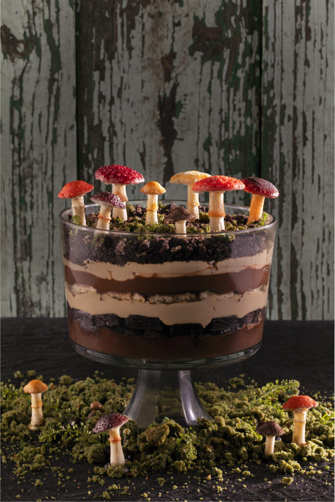 A Girl Scout Cookie Trifle layered in a clear bowl decorated with green cake moss and mushrooms on top and below the glass bowl.