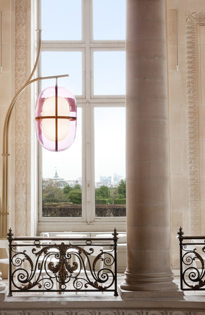 A lamp with a glass pink shell sits in a room with tall reaching windows and columns.