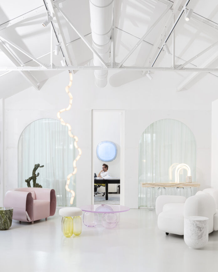 An interior white space with various funky furnishings around the room such as a pink chair on the left side and a blue orb in the middle that designer Mathieu Lehanneur sits beneath.