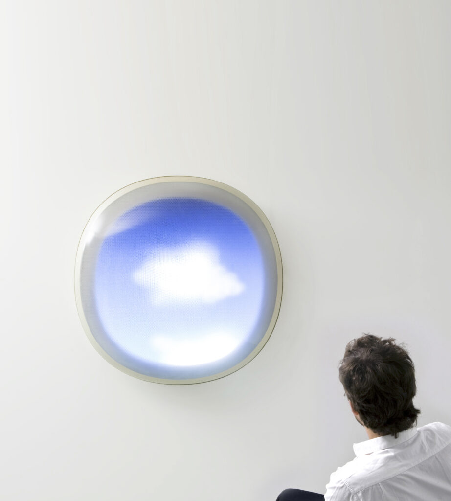 A circle window showing a blue sky and clouds sits on a white wall as Mathieu Lehanneur sits to the right, looking at it.