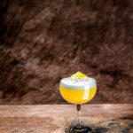 A glass of Mezcal Spring Piña Sur with foam on top, garnished with lemon peel sits on a table against a brown background.