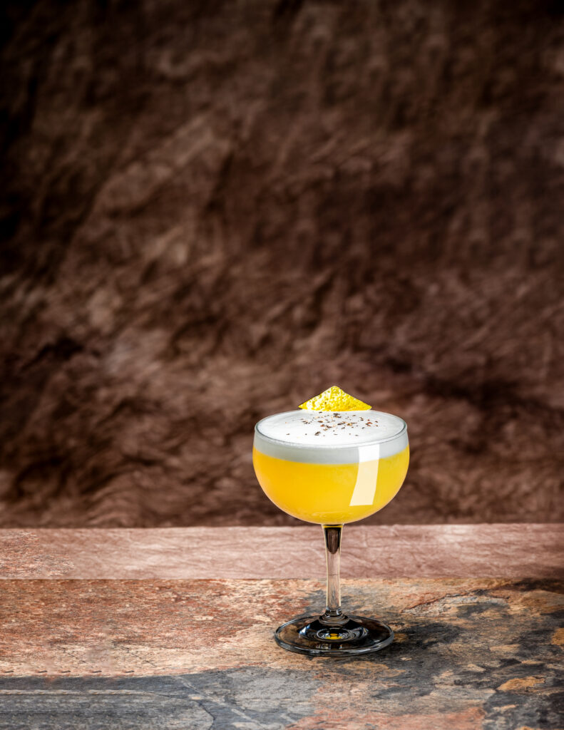 A glass of Mezcal Spring Piña Sur with foam on top, garnished with lemon peel sits on a table against a brown background.