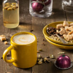 A yellow mug with a latte surrounded by a small glass jar of simple syrup, Nespresso coffee pods, sugar cubes on a little stack of yellow plates and a silver spoon on a wooden surface.