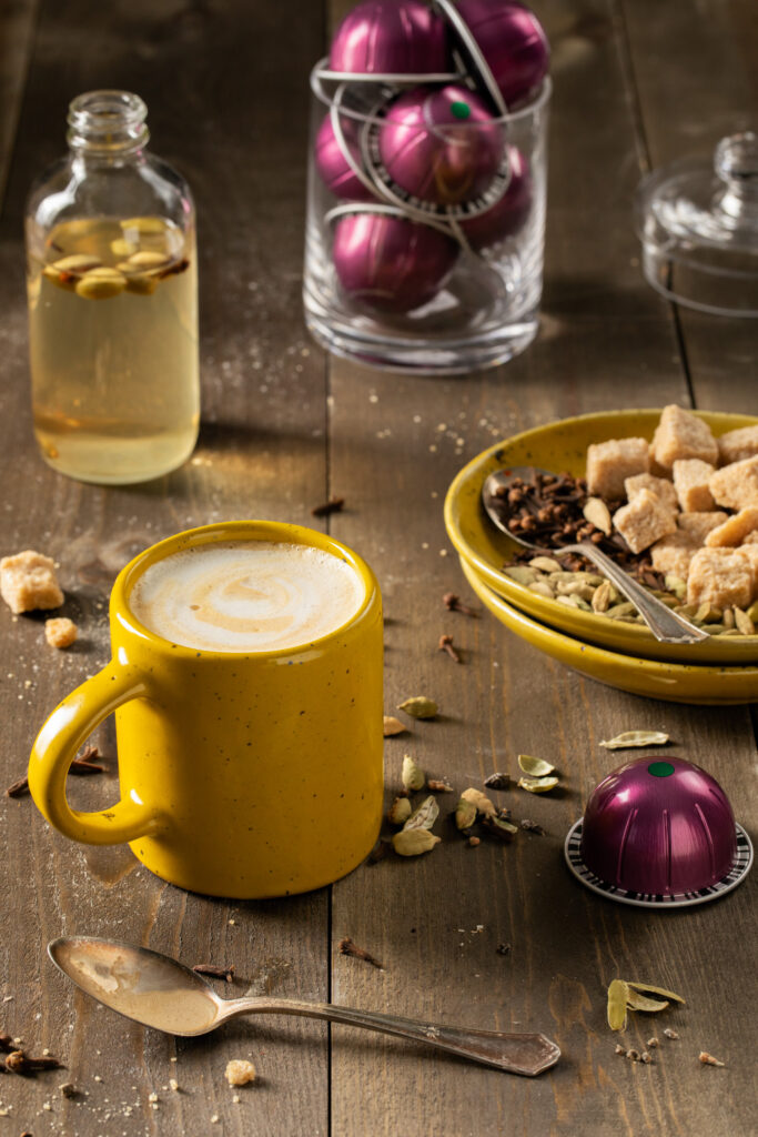 A yellow mug with a latte surrounded by a small glass jar of simple syrup, Nespresso coffee pods, sugar cubes on a little stack of yellow plates and a silver spoon on a wooden surface.