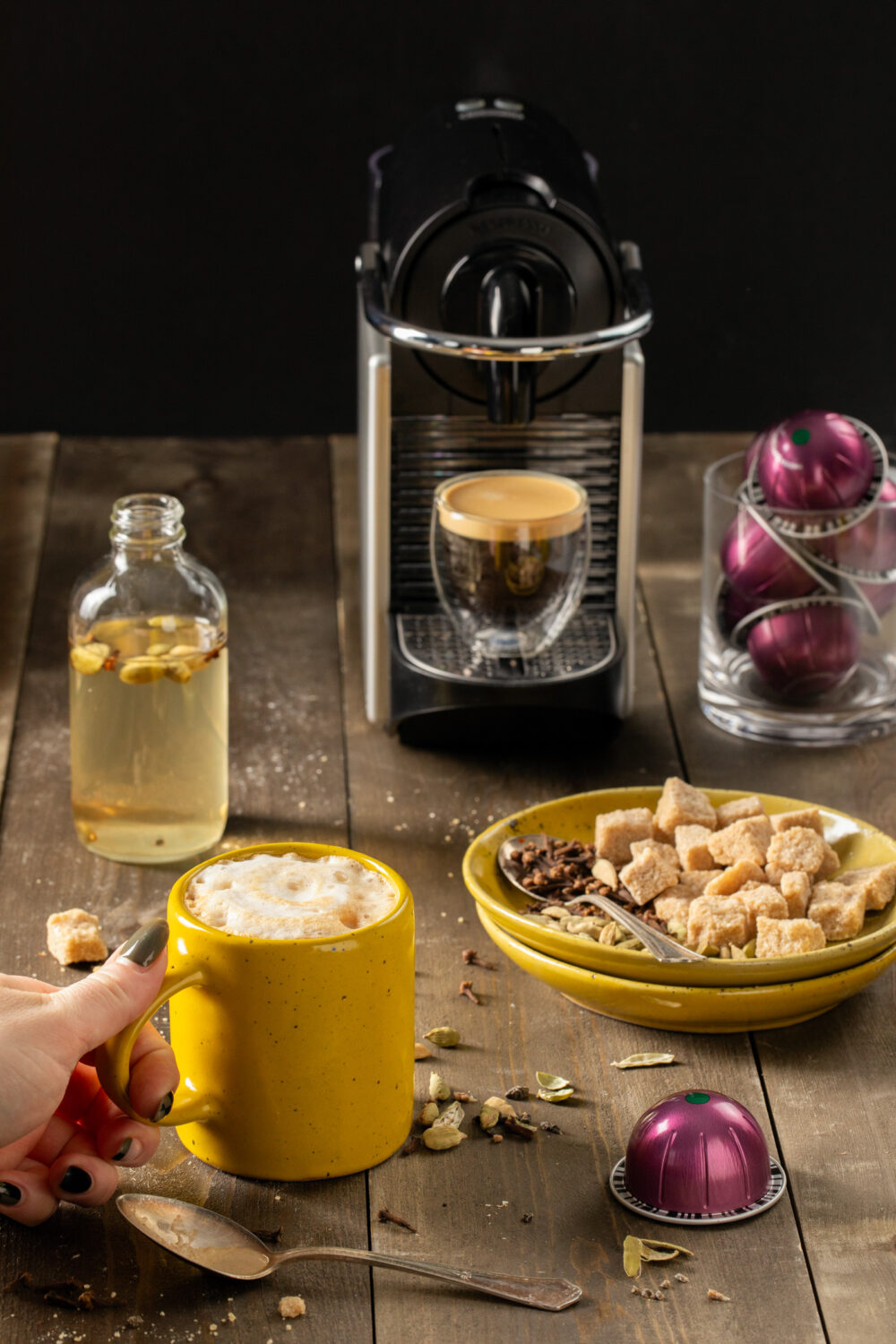 A latte in a yellow cup sits amongst various ingredients and a nespresso machine brewing a shot of espresso. 