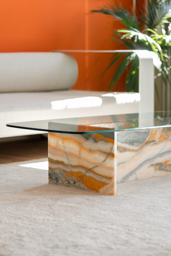 A table with a glass top and orange marble bottom sits in a living room with an orange wall and white couch.