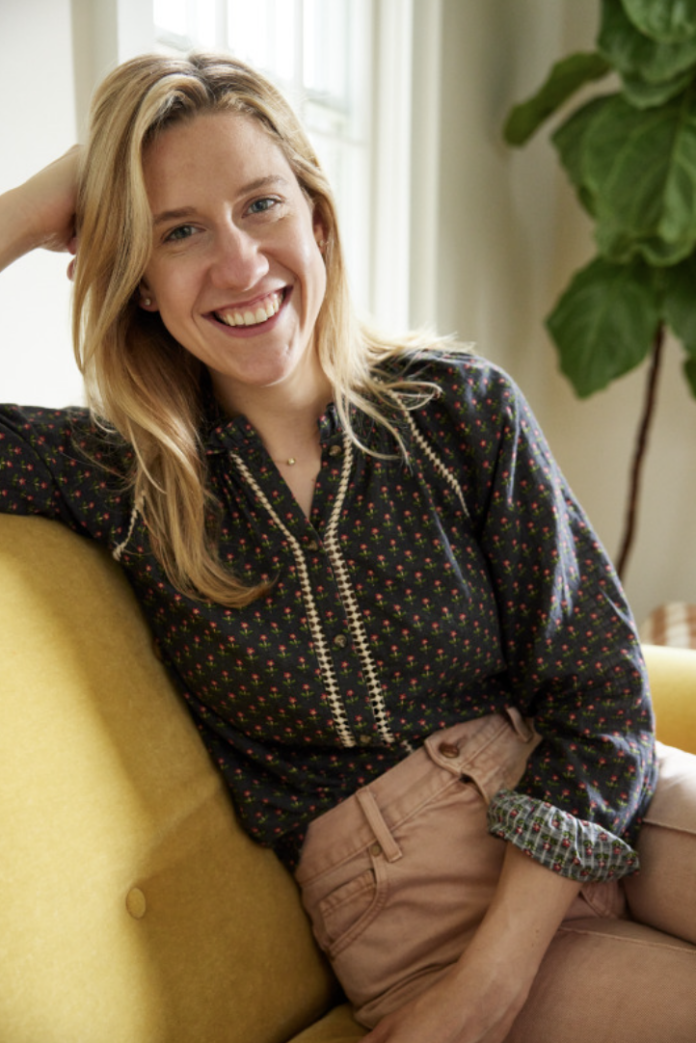 Lidey Heuck smiles on a yellow sofa in her home with her head rests on her fist and a black blouse on.