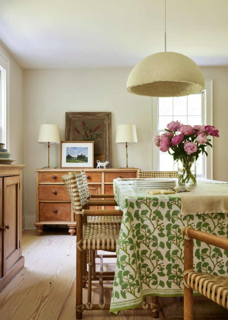 An interior kitchen with beige walls, a wooden dresser in the back, and a table with a green printed tablecloth.