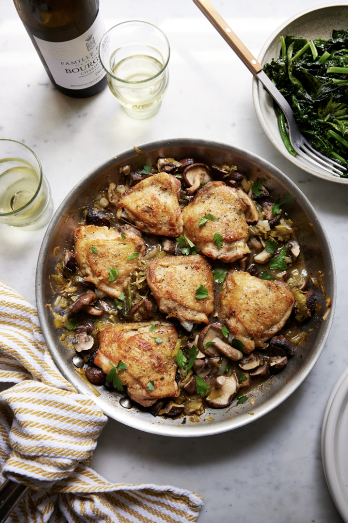 A plate full of crispy chicken thighs in leeks and mushrooms with wine and a salad nearby.