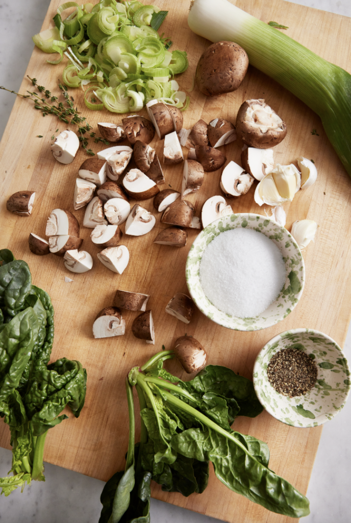 A cutting board holds chopped up mushrooms and leeks with salt and pepper sitting in little bowls.