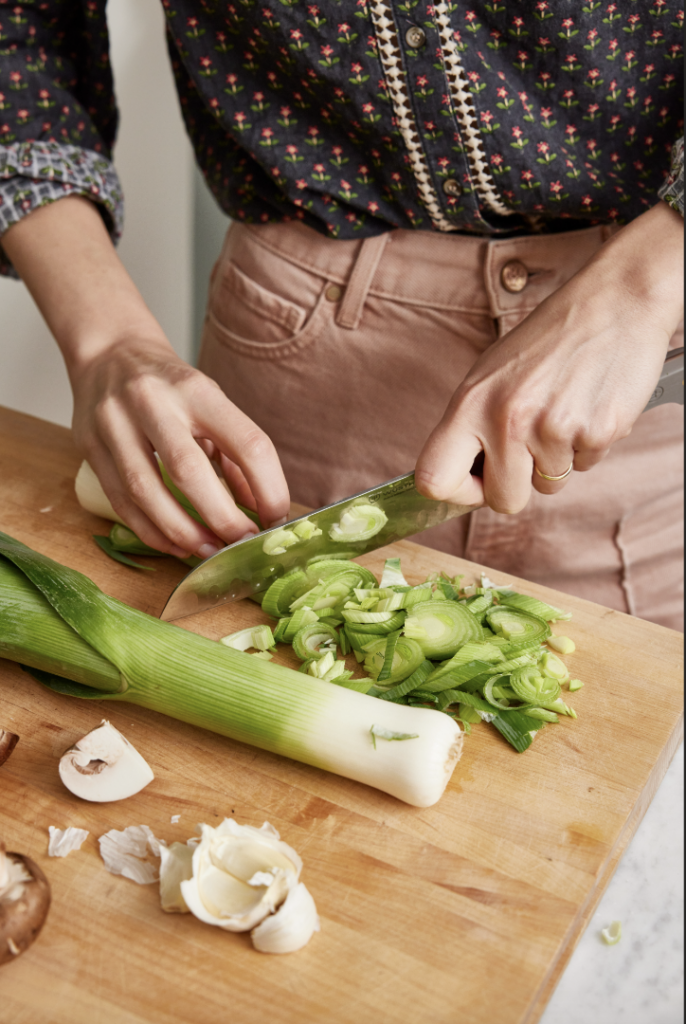 A woman chops up a leek on a wooden cutting board with a few mushrooms nearby.