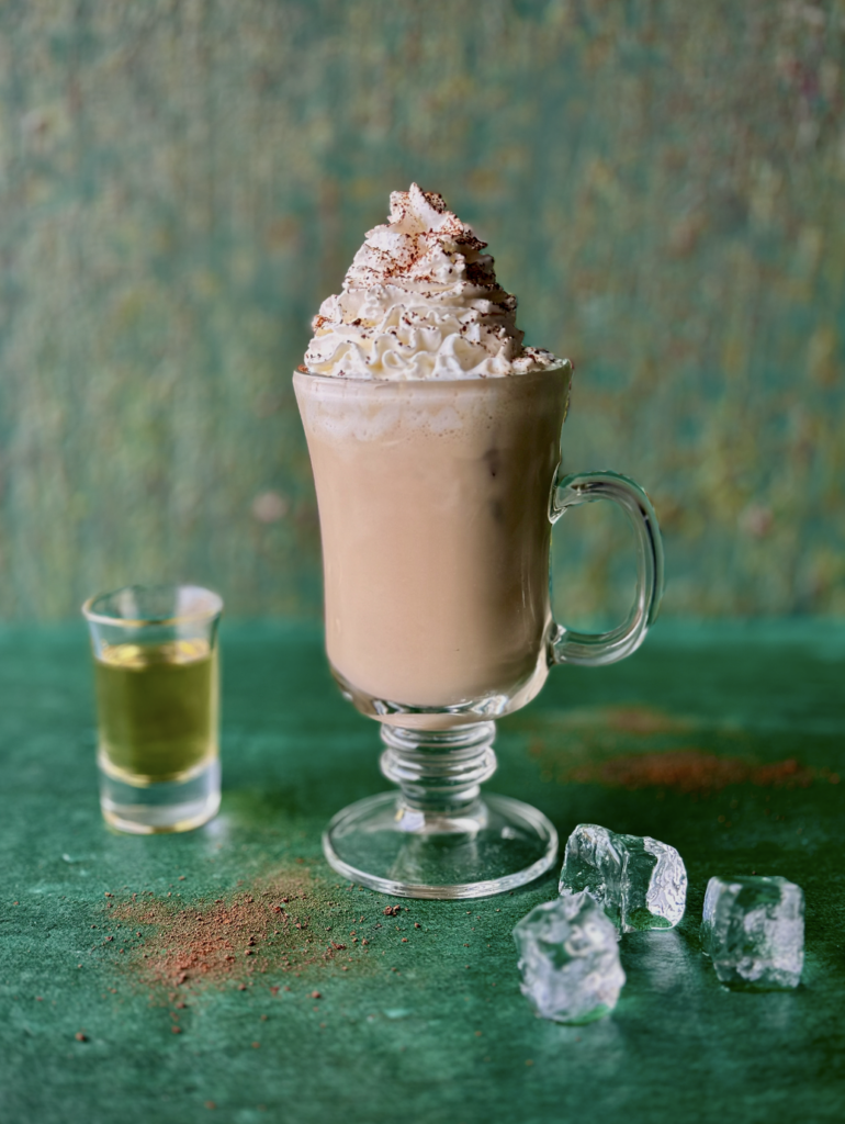 A glass holds an Irish Cream with whipped cream on top, a shot of whiskey to the left, and ice cubes sitting in front of the glass all on a green background.
