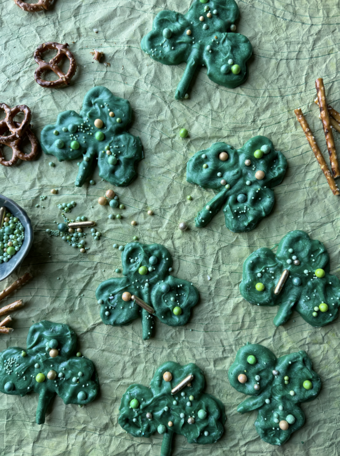Chocolate Pretzel Shamrocks Green Chocolate Pretzel Shamrocks lay on a green textured background with regular pretzels and sprinkles scattered throughout.