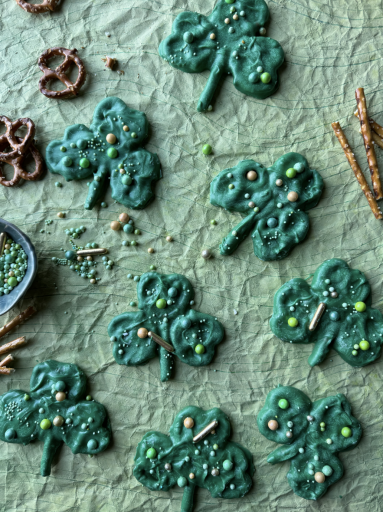 Green Chocolate Pretzel Shamrocks lay on a green textured background with regular pretzels and sprinkles scattered throughout.
