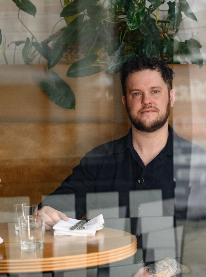 The Vandal's Joey Hilty Talks Family and Food Chef Joey Hilty sits at a table with a reflection in the window framing him.