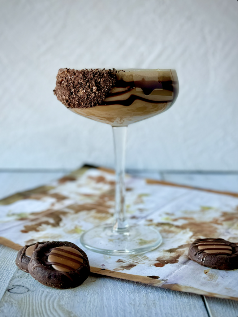 An Adventurefuls® Cocktail is placed in a cocktail glass drizzled with chocolate syrup and rim coated in crushed Adventurefuls® cookies. Three Adventurefuls® cookies sit in front of the glass.