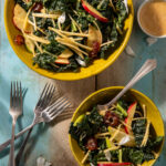 Two yellow bowls hold an an Apple and Rutabaga Salad with a Date Vinaigrette in a small dressing bowl to the top right and forks in the bottom left corner.
