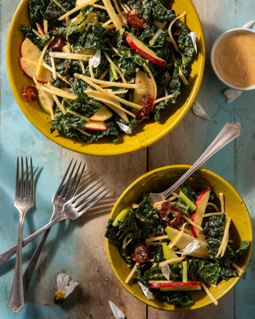 Two yellow bowls hold an an Apple and Rutabaga Salad with a Date Vinaigrette in a small dressing bowl to the top right and forks in the bottom left corner.