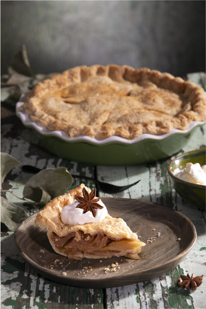 A baked pie sits in a pie tin with a slice of apple pie sitting on a brown plate in front of the pie, topped with an anise star.
