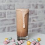 A tall glass with a brown chocolaty drink, foaming over the side, with little pastel colored chocolate Easter eggs on a white surface. Cottontail Cocktail