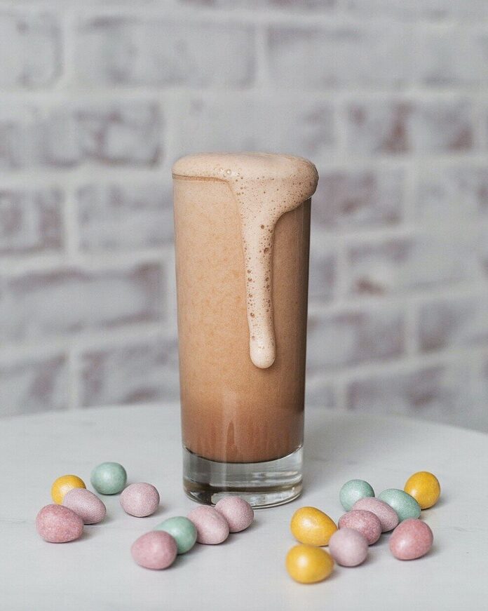 A tall glass with a brown chocolaty drink, foaming over the side, with little pastel colored chocolate Easter eggs on a white surface. Cottontail Cocktail